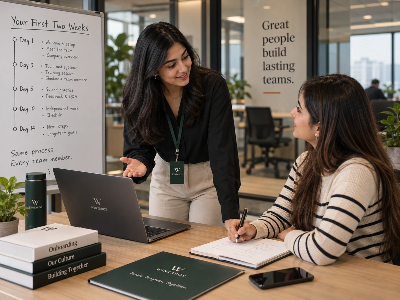 A Wintaros colleague explaining the first two weeks to a new team member, beside a whiteboard that sets out the onboarding plan from day one to day fourteen.