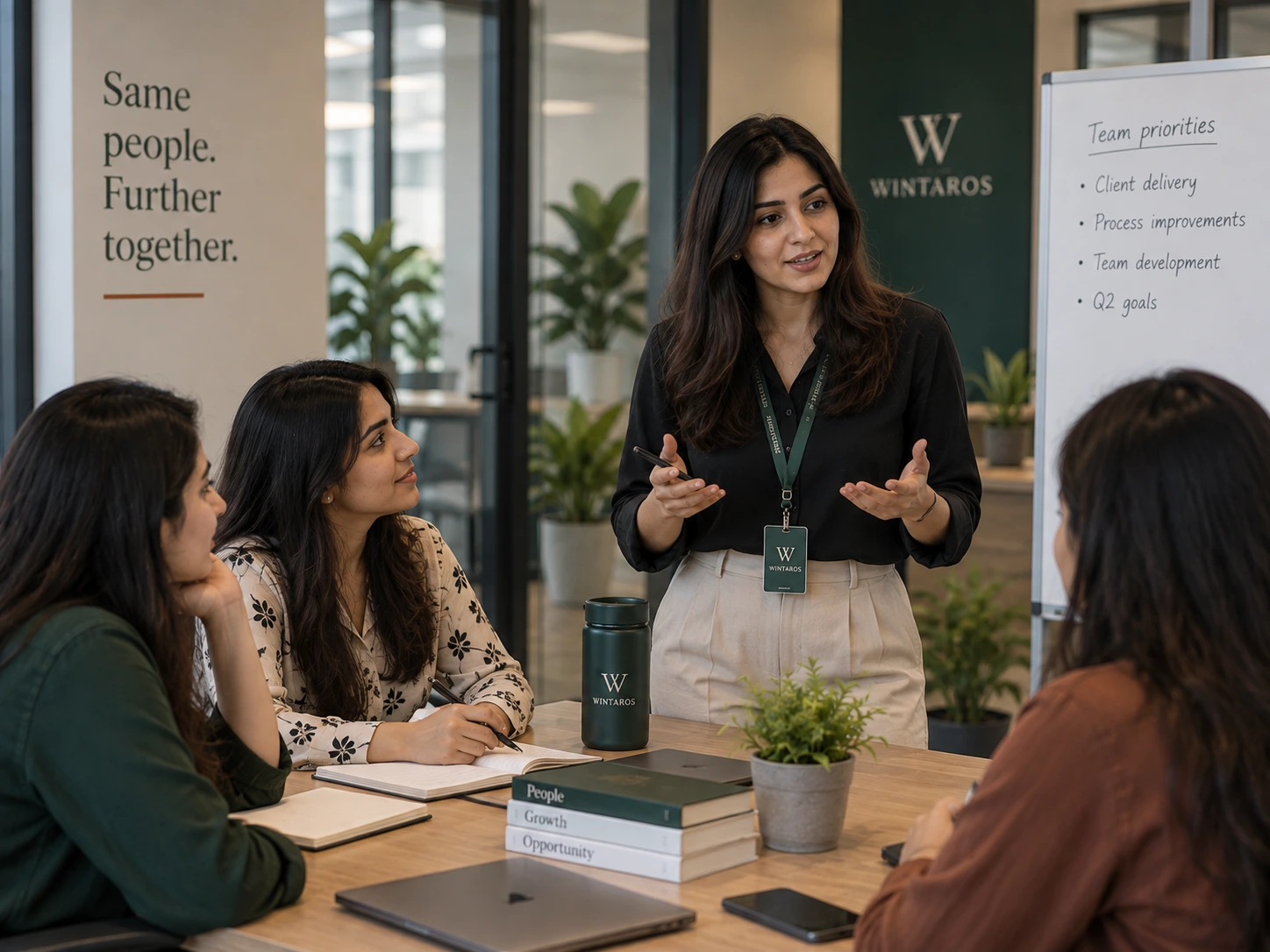 A team lead standing and speaking to three seated colleagues in a meeting room, beside a whiteboard headed &lsquo;Team priorities&rsquo;.