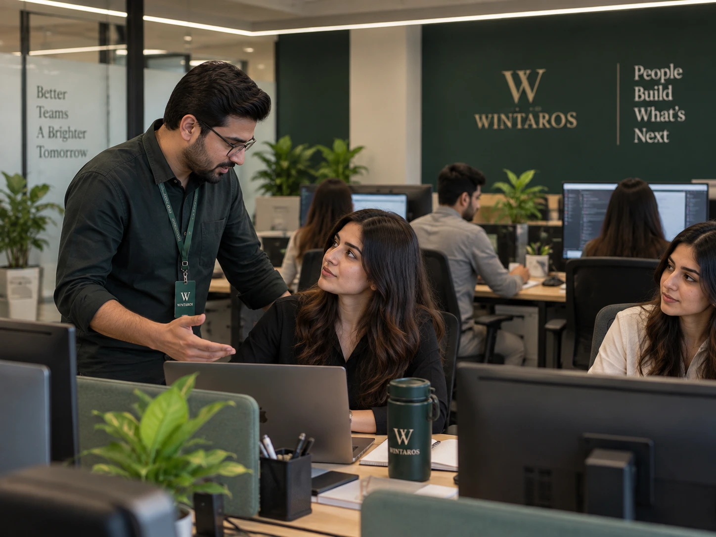 A Wintaros team lead leaning in to talk with a seated colleague at her desk on the office floor, with the Wintaros name on the wall behind.
