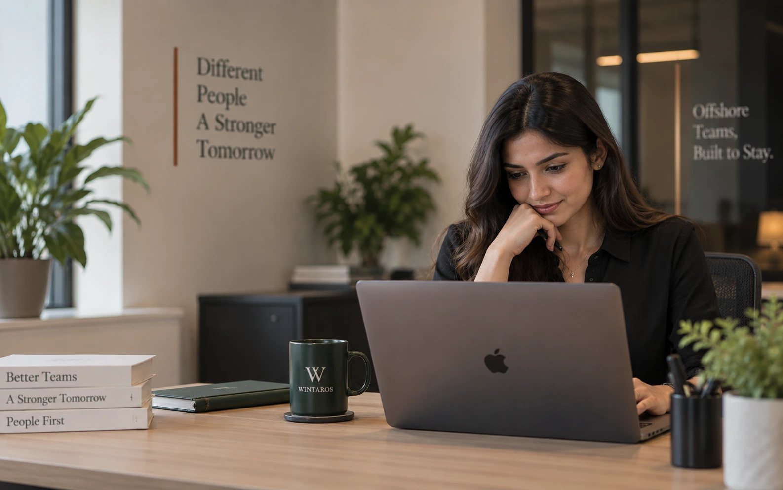 A Wintaros team member reading at her laptop at a tidy desk, with a Wintaros mug beside her and the words &lsquo;Offshore Teams, Built to Stay.&rsquo; on the glass behind.